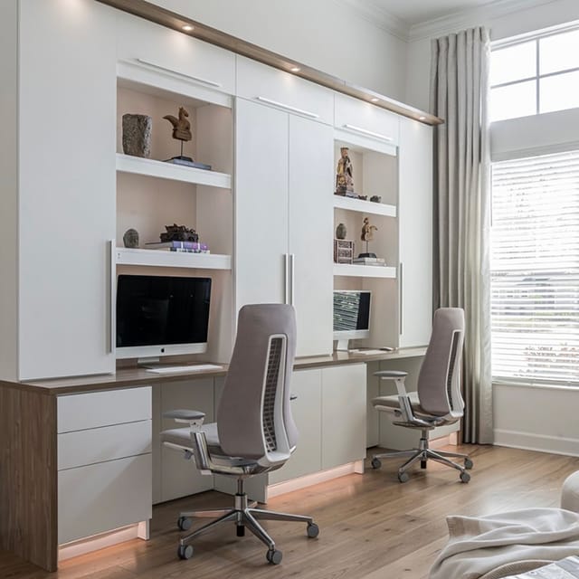 A bright and modern home office featuring two computer workstations, ergonomic chairs, and built-in white shelves and cabinets. The space is illuminated by natural light from a large window with blinds and curtains.